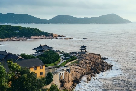 Traditional Chinese black tile temple buildings with yellow walls perched on coastal rocky cliffs, calm sea and green distant mountains at Putuoshan Island