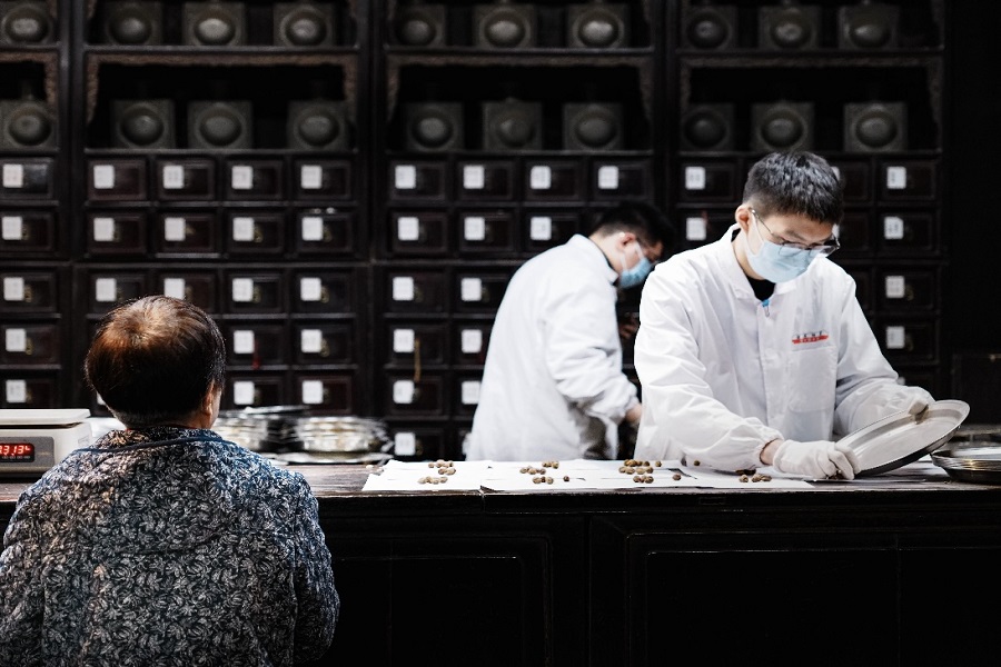 Professional TCM pharmacists in white lab coats weighing medicinal herbs for customers in front of ancient wooden medicine cabinets inside Hu Qing Yu Tang pharmacy