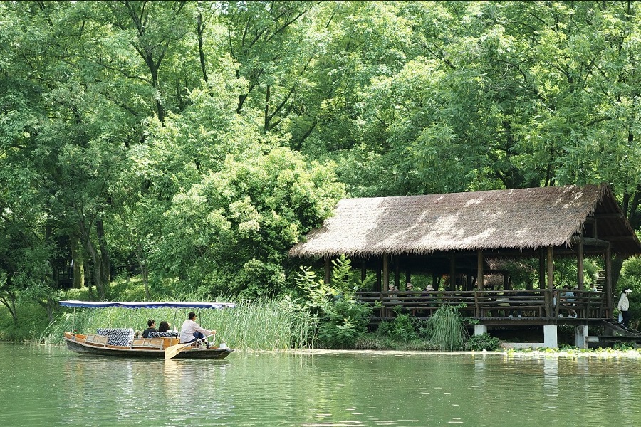 Tourists taking a wooden rowing boat on calm lake surrounded by dense green trees and thatched pavilion by the shore