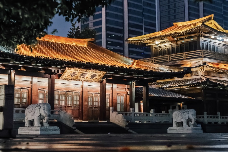 Lit ancient wooden halls of Xiangji Temple with stone elephant sculptures at the gate, classic Chinese Buddhist architecture against modern city buildings at dusk