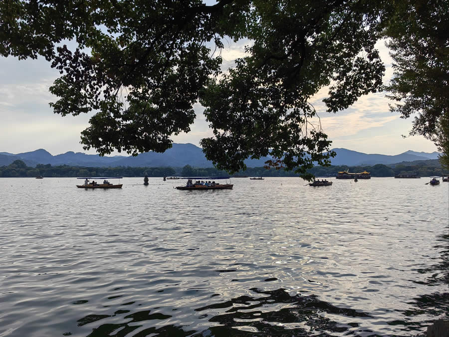 West Lake boating in Hangzhou featuring traditional wooden boats drifting on lake water surrounded by rolling green hills and classical lakeside scenery