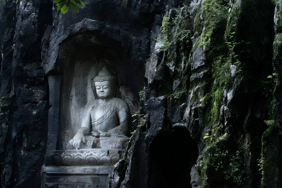 Weathered stone Buddha sculpture in Feilai Peak grotto, carved deep inside rugged dark cliff, green moss and wild plants covering rock walls, retaining primitive solemn charm of Southern Song Buddhist stone art